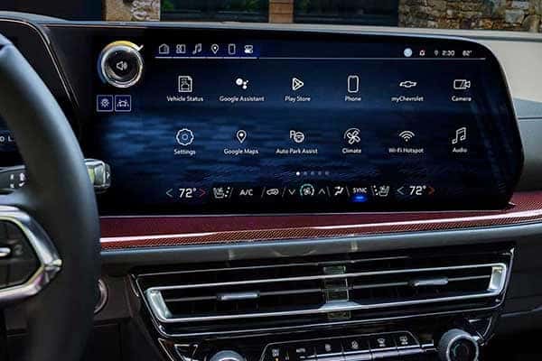 Close-up of a car dashboard featuring a large touchscreen display. The screen shows various icons for vehicle status, Google Assistant, Play Store, phone, camera, and settings. Temperature controls are visible below the screen. Steering wheel partially shown.
