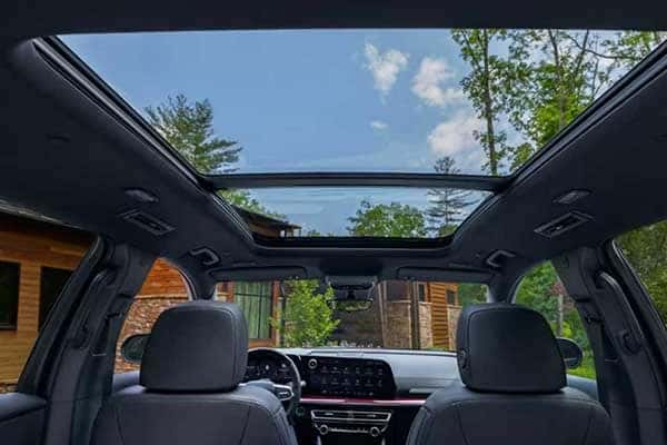 View from inside a car showing the open panoramic sunroof on a sunny day. Trees and a wooden house are visible outside. The car interior features two front seats and a dashboard with a digital display.