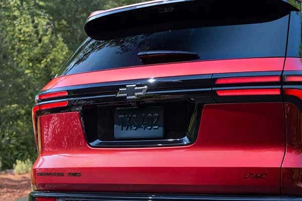 Rear view of a red Chevrolet SUV with a visible bowtie logo. The vehicle features tinted windows and its license plate reads 7K3 402. It is parked outdoors with greenery in the background.
