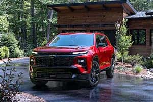 A red SUV is parked in front of a modern wooden house surrounded by trees. The driveway is wet, reflecting the vehicle and greenery. The SUV has a sleek design with black trim and dark wheels.