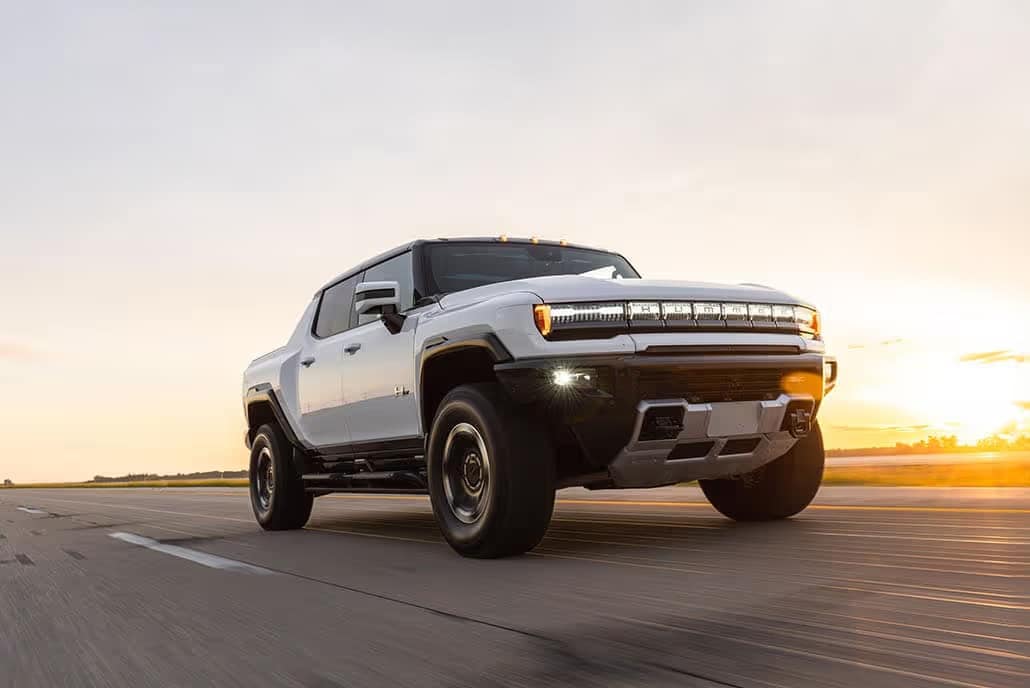 A white pickup truck drives on a road against a backdrop of a sunset sky. The vehicle is large with bright front lights and rugged tires, highlighting an adventurous and powerful design. The road stretches ahead, with a horizon in the distance.