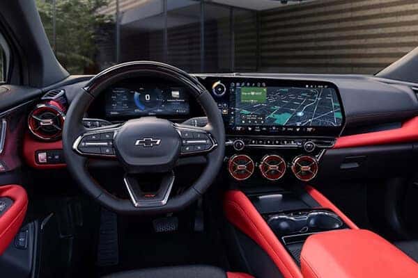 Interior of a car featuring a Chevrolet logo on the steering wheel, a digital dashboard, and a large infotainment screen displaying a map. The interior has red accents and metallic finishes, with three circular air vents below the screen.