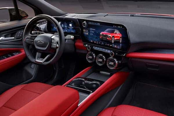 Car interior with red and black seats and dashboard. Features include a large touchscreen display showing a red car, a digital instrument cluster, and three circular air vents. The steering wheel displays a Chevy logo.