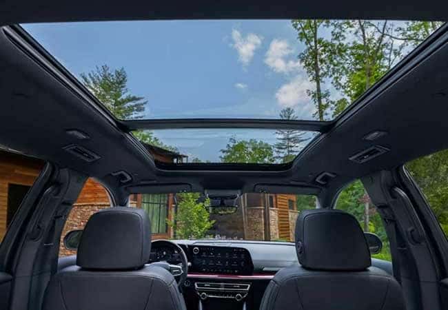 View from the backseat of a car with a panoramic sunroof open to a clear blue sky and surrounding trees. The interior includes two front seats and a modern dashboard. A wooden cabin is visible through the windshield.