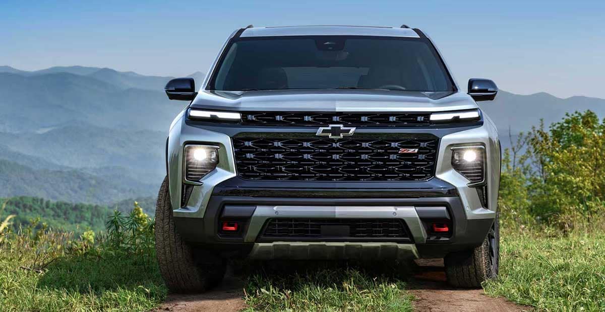Front view of a silver Chevrolet SUV with the brand logo on the grille. It is parked on a dirt path in a mountainous landscape, under a clear blue sky. The headlights are on, and lush greenery surrounds the vehicle.