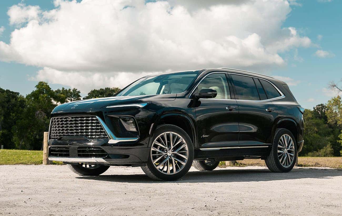 A sleek black SUV parked on a gravel surface under a partly cloudy sky. The vehicle has a modern design with a distinctive grille and shiny alloy wheels. Trees and grass are visible in the background.