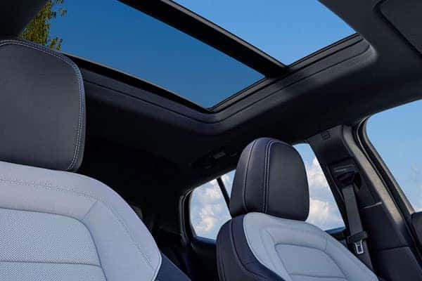 View from inside a car showing two seats and an open sunroof against a clear blue sky. The seats are upholstered in light and dark fabric, and the sunroof provides a wide view of the sky and tree leaves.