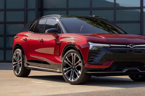 A sleek, modern red SUV with black accents is parked in front of a building with large windows. The vehicle features stylish headlights, a prominent grille, and shiny alloy wheels, showcasing a contemporary automotive design.