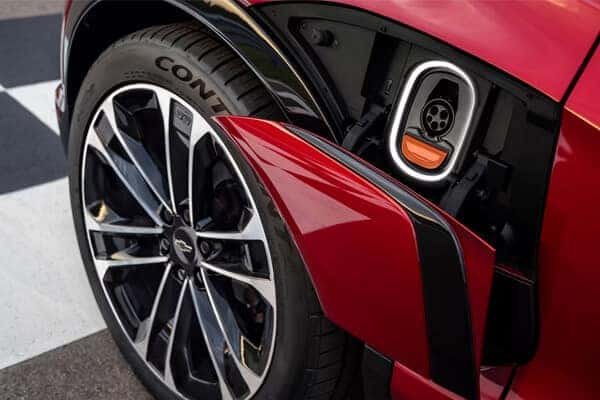 Close-up of a red electric vehicles rear wheel and charging port. The tire has Conti branding, and the port is open, showing a circular socket with an orange latch. The car is parked on a patterned surface.