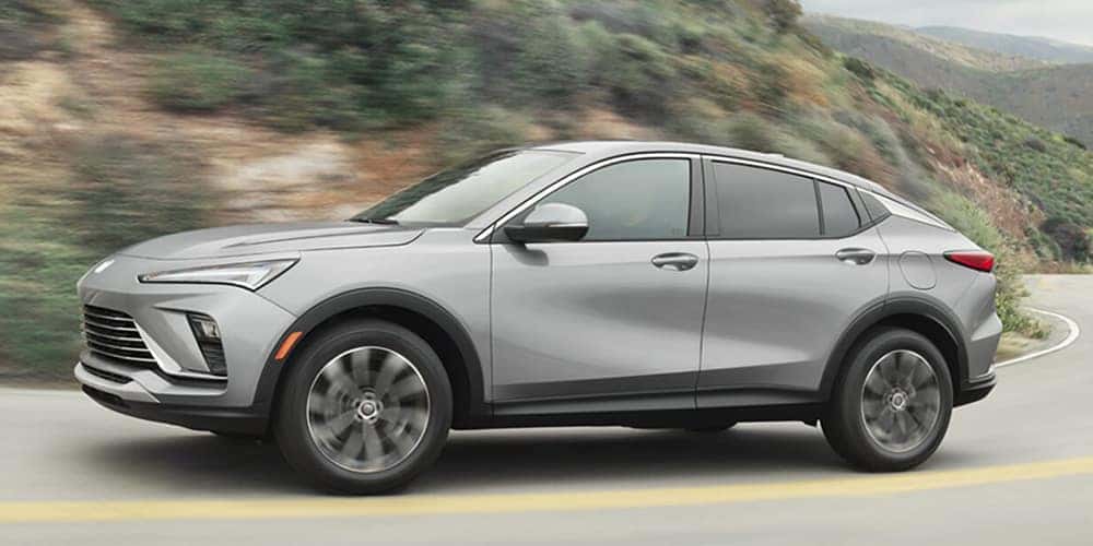 A silver crossover SUV driving on a curved road through a hilly landscape with greenery in the background. The car has sleek lines and a modern design, shown from a side angle.