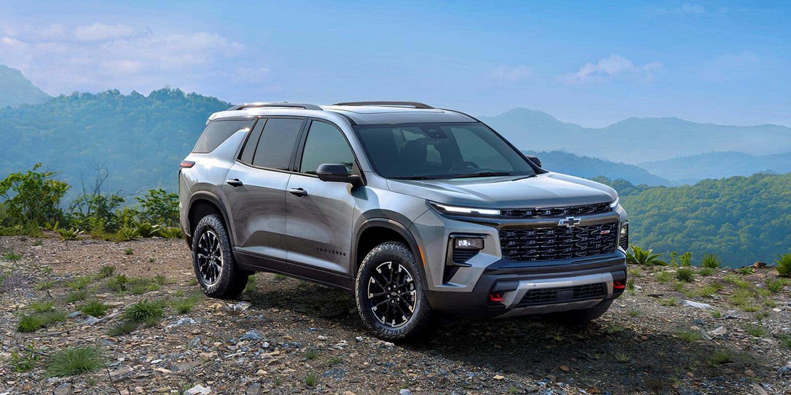 Silver SUV parked on rocky terrain with greenery and mountain range in the background. The SUV is facing slightly to the right, showcasing its front and side profile. The sky is clear and blue.