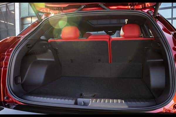 Open trunk of a red SUV with red interior seats visible. The seats can fold down for more cargo space. Sunlight creates lens flare on the left. The surrounding area includes some industrial buildings.