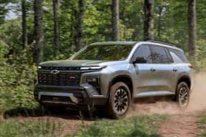 A silver SUV driving on a dirt path through a forest, surrounded by green trees and foliage. Dust is visible behind the vehicle, indicating motion. The vehicles front is prominently shown with a visible grille and headlights.