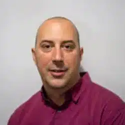 Bald man smiling in a magenta shirt against a neutral background.