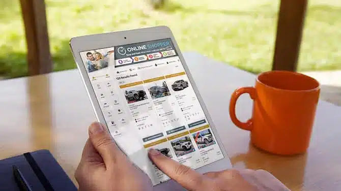 Person browsing car listings on a tablet with an orange coffee mug nearby.