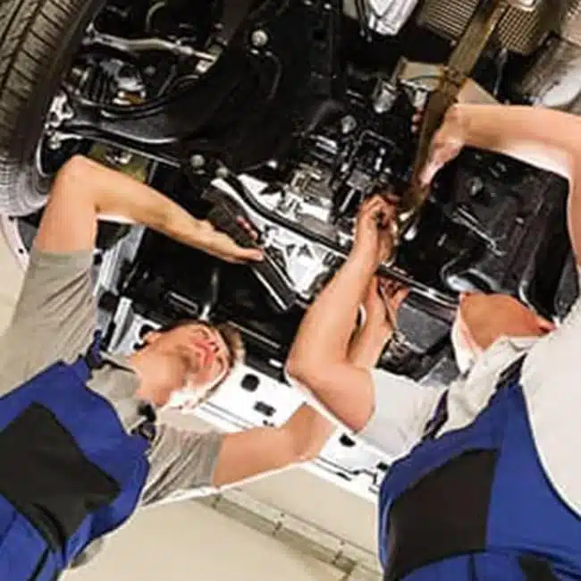 Mechanics in blue uniforms repairing a car's underside in an auto repair shop.
