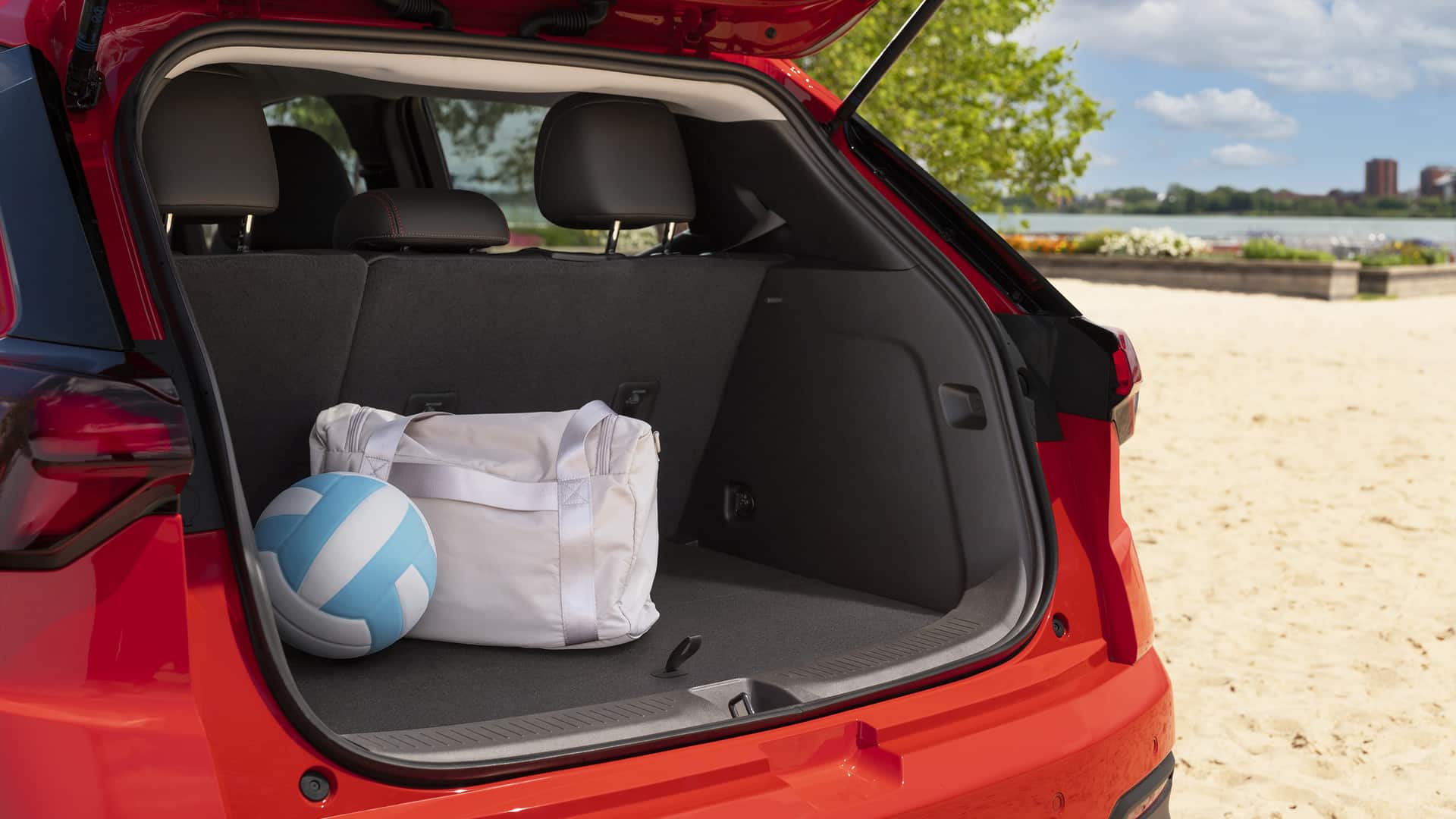 Open trunk of a red car with a white duffel bag and a blue volleyball at a sunny beach.
