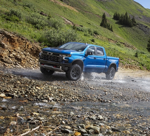 Blue pickup truck driving through a rocky stream in a lush, mountainous landscape.