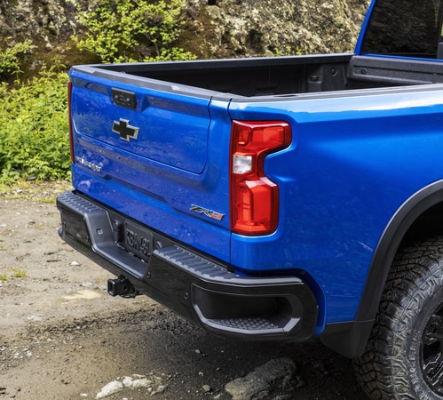 Blue pickup truck with an open bed parked on a dirt road, surrounded by greenery and rocky terrain.