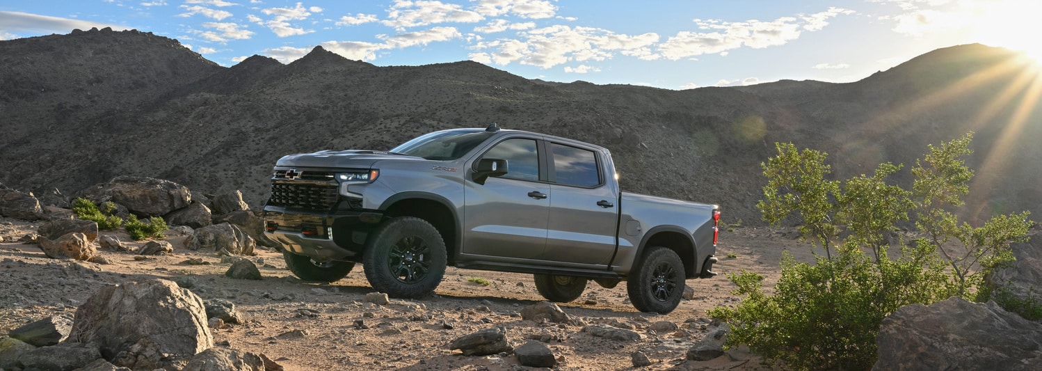 A shiny red SUV is parked in a dimly lit urban setting at night. The car features a sleek design with distinctive wheel rims, and illuminated headlights, reflecting light on the wet pavement. Modern architectural elements are visible in the background.