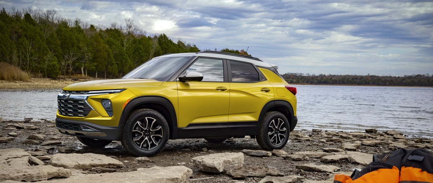 A green SUV is parked on a paved surface near a calm body of water. The car features a prominent grille and sleek design. In the background, hills are visible under a clear blue sky.