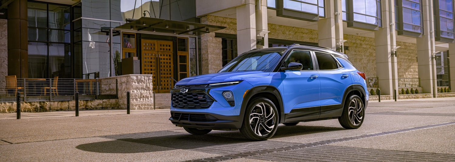 A shiny red SUV is parked in a dimly lit urban setting at night. The car features a sleek design with distinctive wheel rims, and illuminated headlights, reflecting light on the wet pavement. Modern architectural elements are visible in the background.