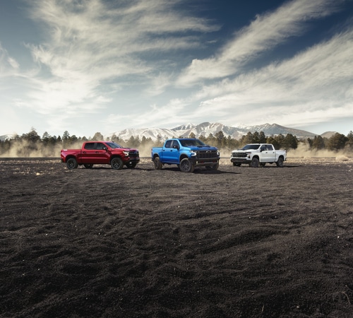 Three trucks—red, blue, and white—parked on a dirt field with mountains in the background.