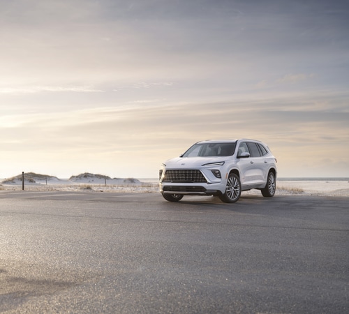 A sleek white SUV parked on a coastal road during a pink and orange sunset, showcasing scenic views.