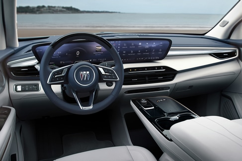View from the backseat of a car showing two people in the front seats. The driver is holding the steering wheel. The sunroof is open, revealing a blue sky and tall buildings outside. The dashboard and rearview mirror are visible.
