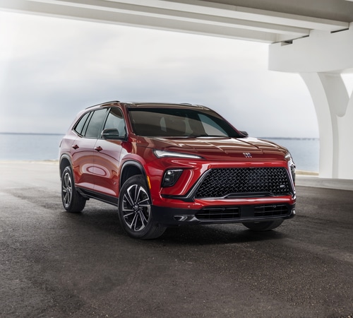 Red SUV parked under a modern structure by the waterfront, featuring a sleek design and prominent grille.
