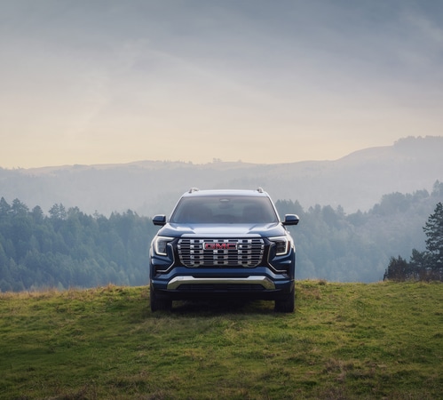 Blue GMC SUV on a grassy hilltop with a misty forest and mountain backdrop.