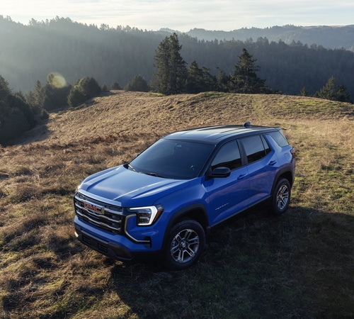 Blue SUV parked on a grassy hilltop with a scenic mountain backdrop under a cloudy sky.