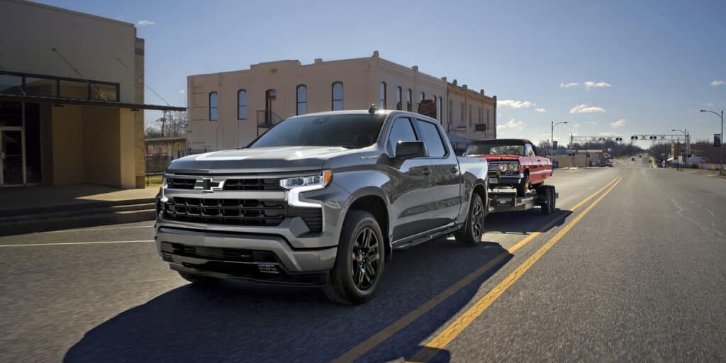 Silver pickup truck towing a red vintage car on an empty urban road under a clear blue sky.