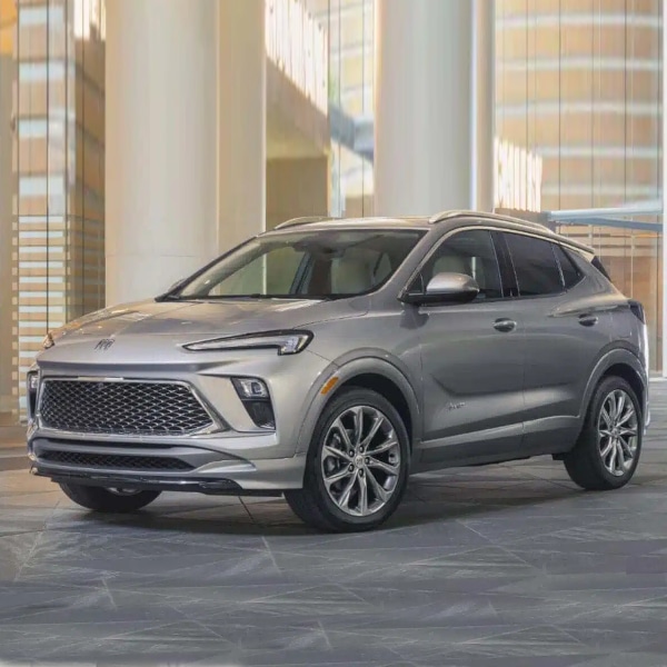 A silver SUV with sleek design and chrome accents is parked indoors on a tiled floor. It has a large grille, sharp headlights, and sporty wheels, set against a modern architectural background with columns and glass.