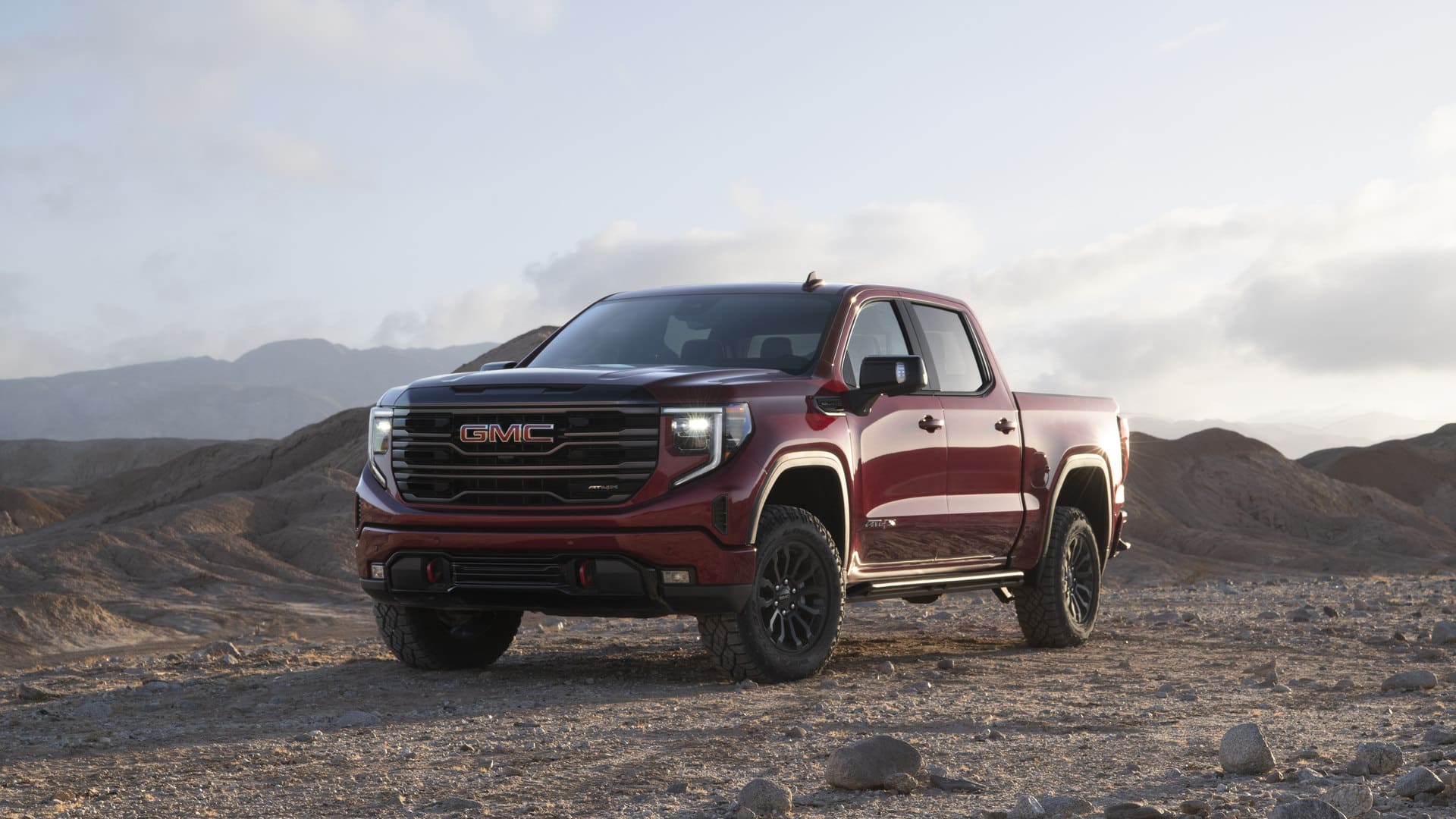 Red GMC truck parked on rocky terrain with hills in the background under a cloudy sky.