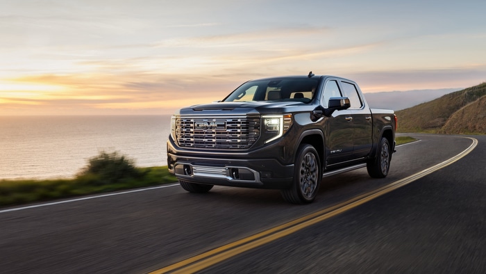 A black pickup truck driving on a scenic coastal road at sunset, with an ocean view in the background.