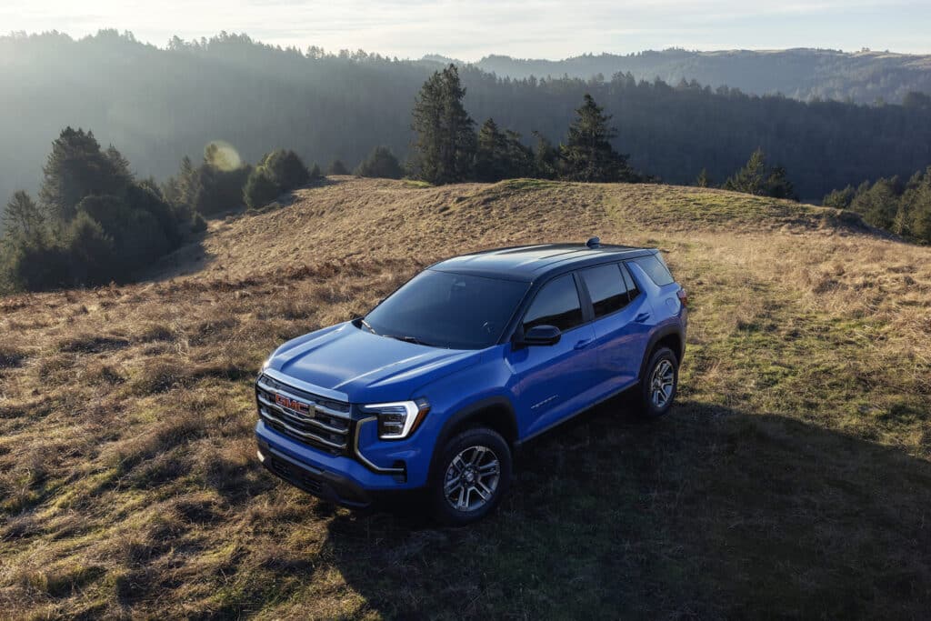 Blue GMC SUV parked on grassy hill with forest scenery under clear sky.