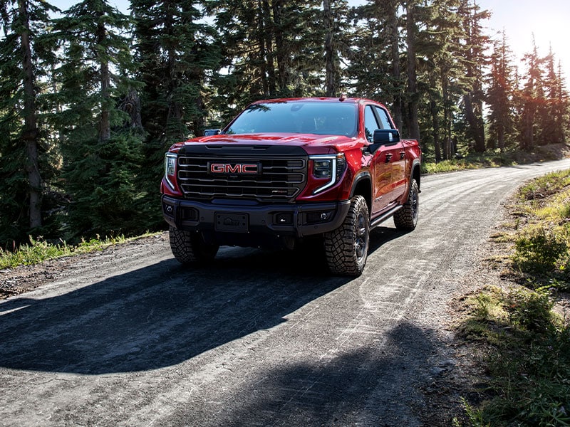 Red GMC pickup truck on a forest road under sunlight, surrounded by tall pine trees.